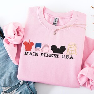 K&ouml;nnte beinhalten: Ein hellrosa Sweatshirt mit gestickten Designs und dem Text "MAIN STREET U.S.A.". Das Design umfasst einen roten Mickey Mouse-Kopf, eine blaue amerikanische Flagge, einen schwarzen Mickey Mouse-Kopf und ein goldfarbenes Bogenfenster.