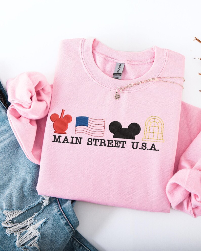 K&ouml;nnte beinhalten: Ein hellrosa Sweatshirt mit gestickten Designs und dem Text "MAIN STREET U.S.A.". Das Design umfasst einen roten Mickey Mouse-Kopf, eine blaue amerikanische Flagge, einen schwarzen Mickey Mouse-Kopf und ein goldfarbenes Bogenfenster.