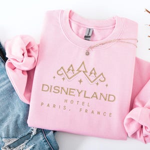 May include: A light pink sweatshirt with the words "Disneyland Hotel Paris, France" in gold lettering. The sweatshirt has a crew neck and long sleeves. A gold necklace with a small charm is draped across the sweatshirt.