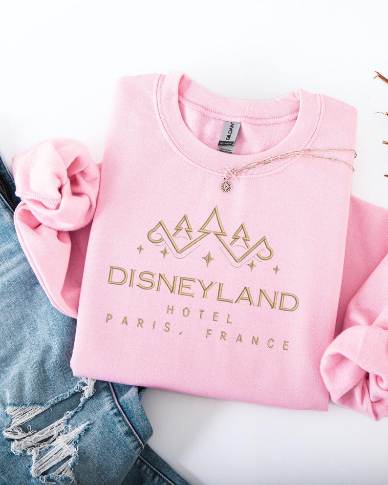 May include: A light pink sweatshirt with the words "Disneyland Hotel Paris, France" in gold lettering. The sweatshirt has a crew neck and long sleeves. A gold necklace with a small charm is draped across the sweatshirt.
