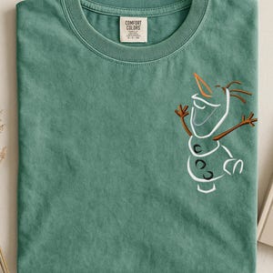 May include: A sage green t-shirt with a crew neck and short sleeves. The shirt features an embroidered outline of Olaf from Frozen on the front. The tag reads "Comfort Colors".