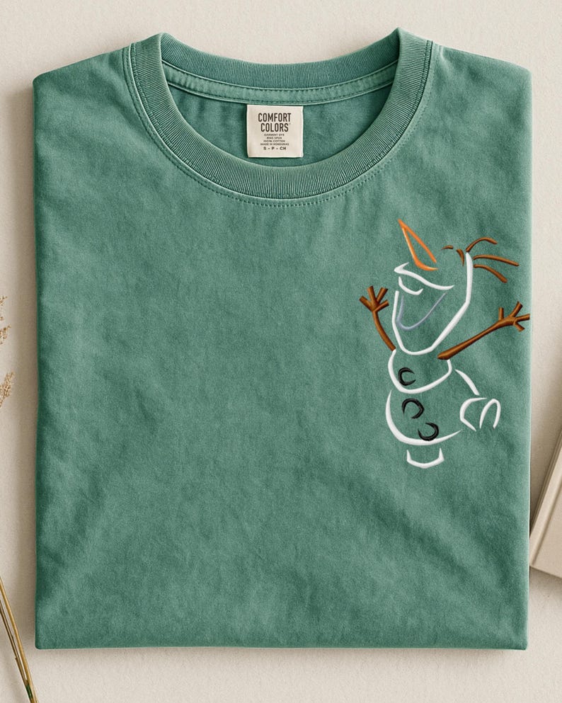 May include: A sage green t-shirt with a crew neck and short sleeves. The shirt features an embroidered outline of Olaf from Frozen on the front. The tag reads "Comfort Colors".
