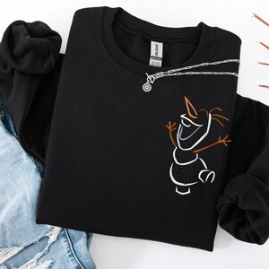 May include: Black crew neck sweatshirt with a white and brown outline of a snowman character. A silver necklace with a circular pendant rests on the sweatshirt. The sweatshirt is folded on blue jeans.