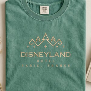 May include: A sage green t-shirt with the words "Disneyland Hotel Paris, France" in a gold-colored font. The shirt features a mountain-like design above the text. The shirt is made by Comfort Colors.