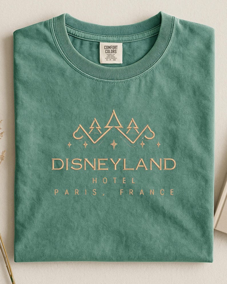 May include: A sage green t-shirt with the words "Disneyland Hotel Paris, France" in a gold-colored font. The shirt features a mountain-like design above the text. The shirt is made by Comfort Colors.