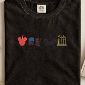 K&ouml;nnte beinhalten: Schwarzes T-Shirt mit gestickten Designs: ein roter Mickey Mouse-Kopf, eine blau-rote Flagge, ein schwarzer Mickey Mouse-Kopf und ein gelbes Bogenfenster. Der Text "MAIN STREET U.S.A." ist darunter gestickt. Mit Comfort Colors-Etikett.