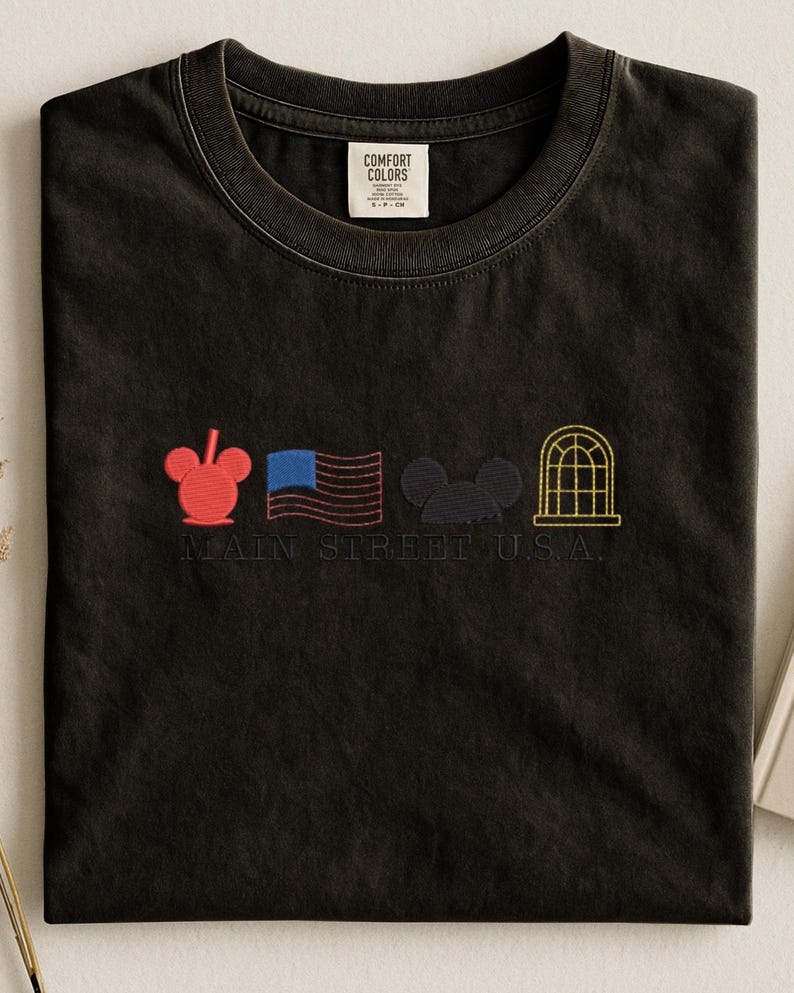 K&ouml;nnte beinhalten: Schwarzes T-Shirt mit gestickten Designs: ein roter Mickey Mouse-Kopf, eine blau-rote Flagge, ein schwarzer Mickey Mouse-Kopf und ein gelbes Bogenfenster. Der Text "MAIN STREET U.S.A." ist darunter gestickt. Mit Comfort Colors-Etikett.