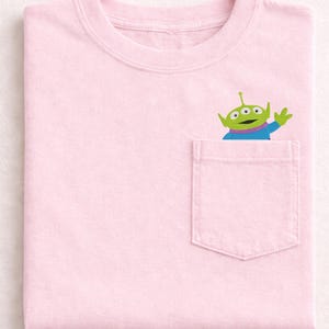 May include: A light pink t-shirt with a pocket featuring a cartoon alien character. The alien is green with three eyes, a purple neck, and a blue shirt, waving from the pocket. The shirt is folded neatly.