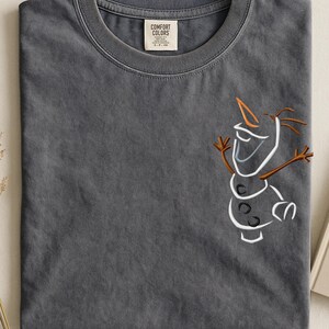 May include: A dark grey t-shirt with an embroidered outline of Olaf from Frozen. The design features white, orange, and brown details. The shirt has a crew neck and a tag that reads "Comfort Colors".