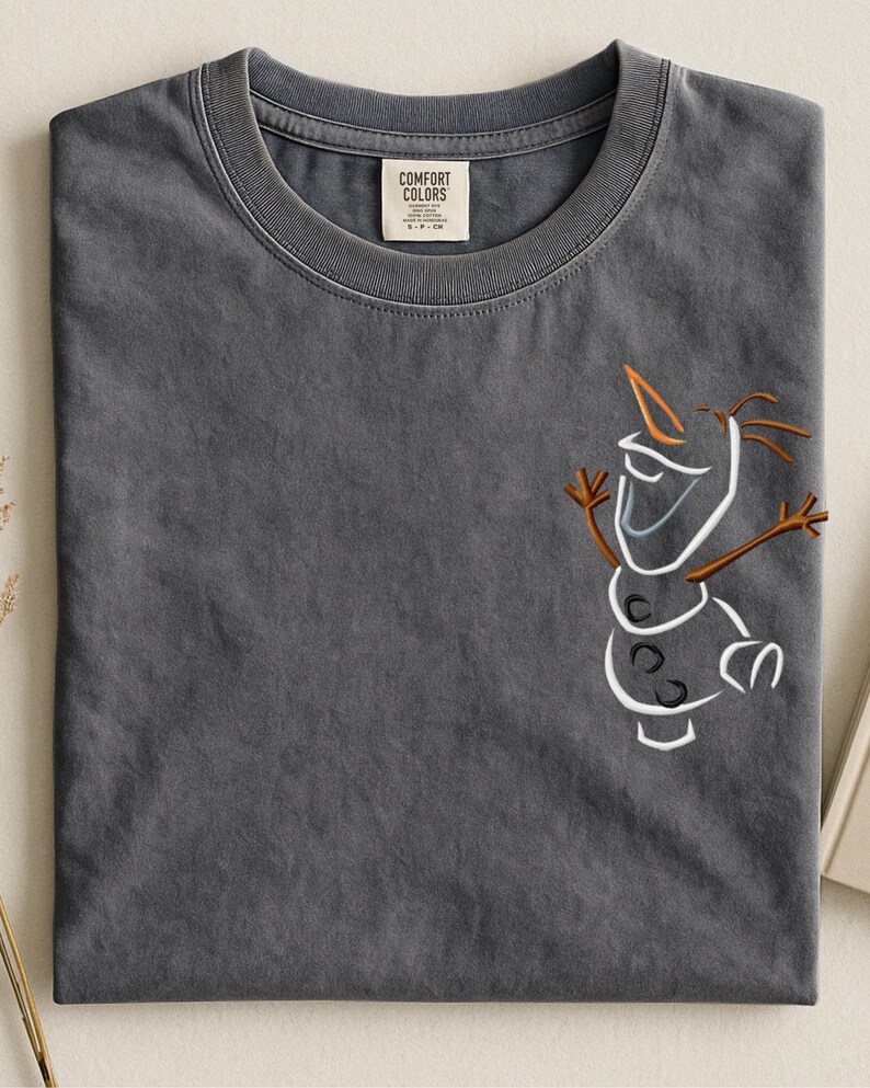 May include: A dark grey t-shirt with an embroidered outline of Olaf from Frozen. The design features white, orange, and brown details. The shirt has a crew neck and a tag that reads "Comfort Colors".