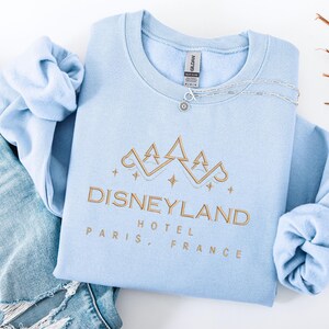 May include: A light blue sweatshirt with the words "Disneyland Hotel Paris, France" embroidered in gold. The sweatshirt has a crew neck and long sleeves. A silver necklace with a small pendant rests on the shirt.
