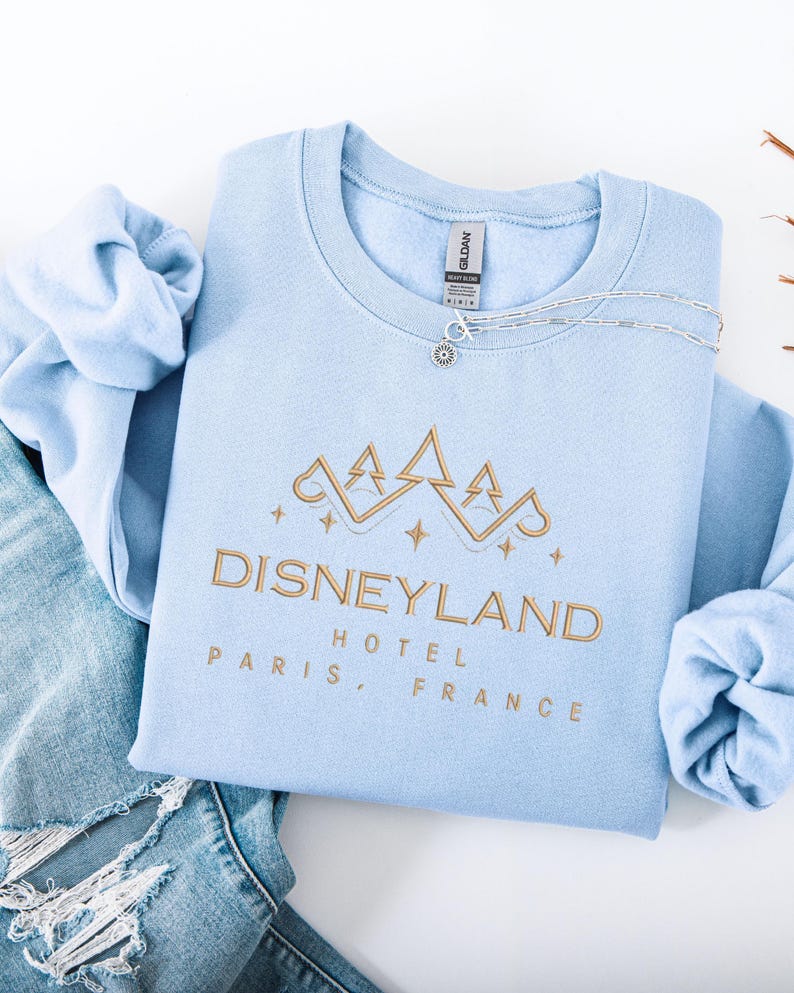 May include: A light blue sweatshirt with the words "Disneyland Hotel Paris, France" embroidered in gold. The sweatshirt has a crew neck and long sleeves. A silver necklace with a small pendant rests on the shirt.