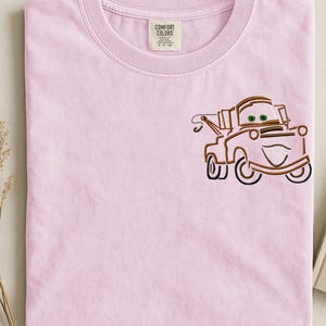 Puede incluir: Una camiseta rosa claro Comfort Colors con un dise&ntilde;o bordado de un cami&oacute;n de dibujos animados. El cami&oacute;n es marr&oacute;n con ojos verdes y una cara sonriente. La camiseta tiene cuello redondo y una etiqueta que dice "Comfort Colors".
