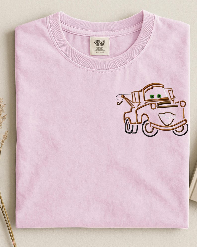 Puede incluir: Una camiseta rosa claro Comfort Colors con un dise&ntilde;o bordado de un cami&oacute;n de dibujos animados. El cami&oacute;n es marr&oacute;n con ojos verdes y una cara sonriente. La camiseta tiene cuello redondo y una etiqueta que dice "Comfort Colors".