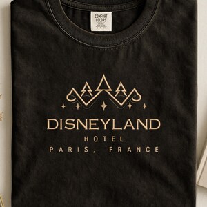 May include: Black t-shirt with a gold-colored Disneyland Hotel Paris, France graphic. The design features a crown and stars above the text. The shirt has a crew neck and a tag that says "Comfort Colors."