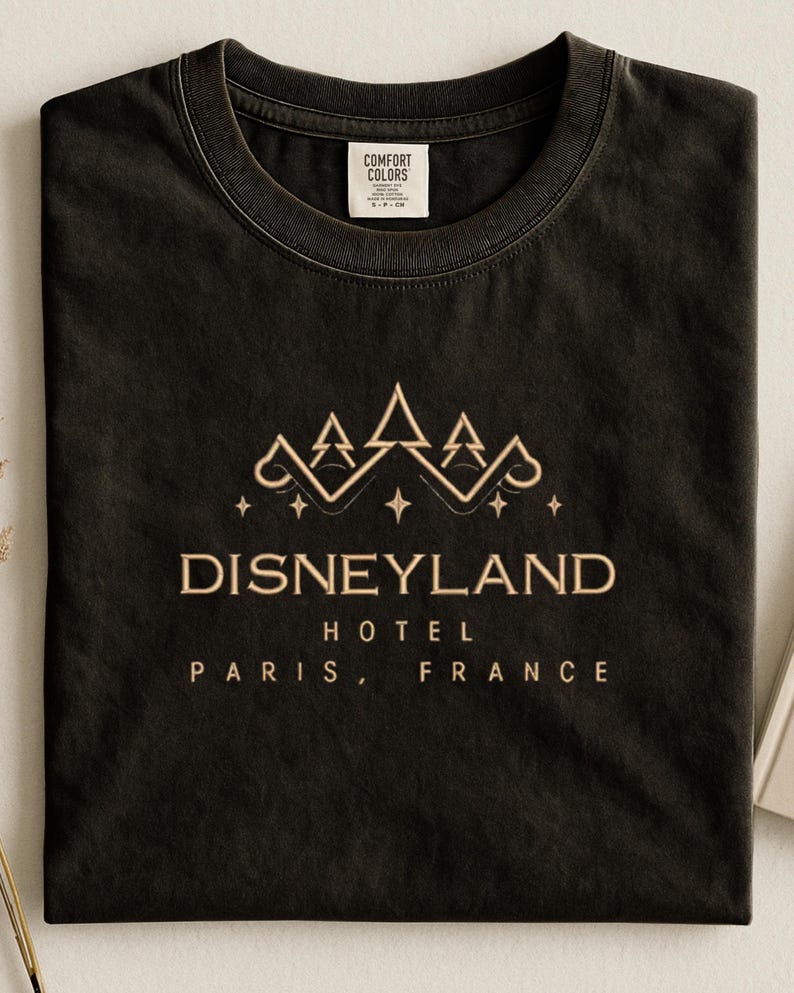May include: Black t-shirt with a gold-colored Disneyland Hotel Paris, France graphic. The design features a crown and stars above the text. The shirt has a crew neck and a tag that says "Comfort Colors."