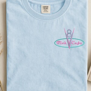 Puede incluir: Camiseta azul claro con el texto bordado "Flo's Cafe" en un dise&ntilde;o retro. El dise&ntilde;o presenta un logotipo "V8" rosa dentro de un &oacute;valo turquesa. La camiseta tiene cuello redondo y una peque&ntilde;a etiqueta en la parte superior.