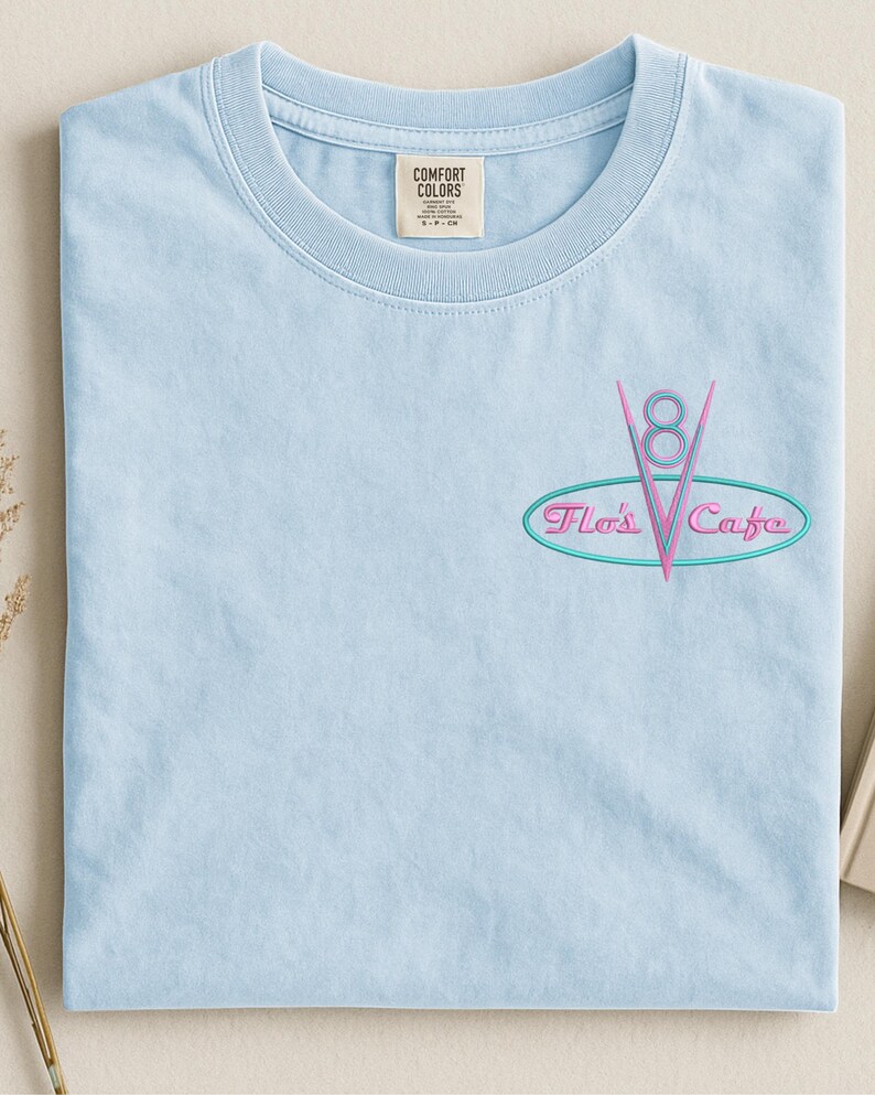Puede incluir: Camiseta azul claro con el texto bordado "Flo's Cafe" en un dise&ntilde;o retro. El dise&ntilde;o presenta un logotipo "V8" rosa dentro de un &oacute;valo turquesa. La camiseta tiene cuello redondo y una peque&ntilde;a etiqueta en la parte superior.