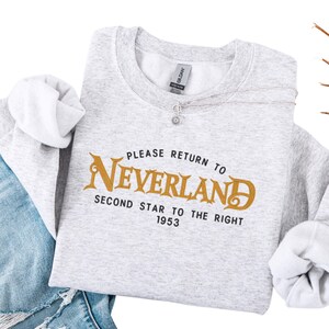 Puede incluir: Sudadera gris claro con el texto "PLEASE RETURN TO NEVERLAND" en letras doradas. Debajo, las palabras "SECOND STAR TO THE RIGHT 1953" están en blanco. La sudadera está doblada sobre unos vaqueros azules.