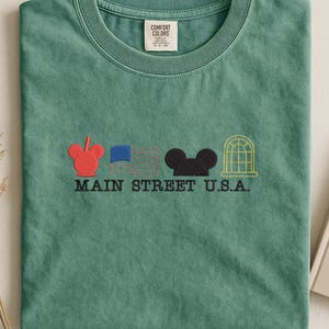 K&ouml;nnte beinhalten: Ein meergr&uuml;nes T-Shirt mit gestickten Designs und dem Text "MAIN STREET U.S.A.". Das Design umfasst einen roten Mickey Mouse-Kopf, eine blaue Flagge, einen schwarzen Mickey Mouse-Kopf und ein gelbes Bogenfenster.