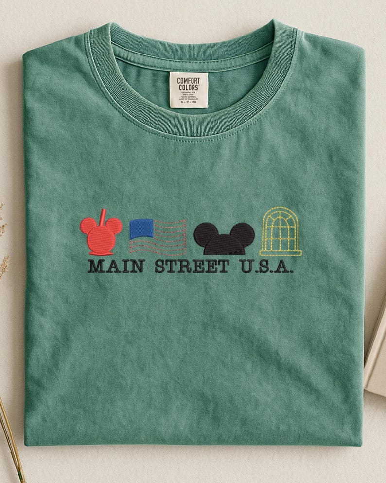 K&ouml;nnte beinhalten: Ein meergr&uuml;nes T-Shirt mit gestickten Designs und dem Text "MAIN STREET U.S.A.". Das Design umfasst einen roten Mickey Mouse-Kopf, eine blaue Flagge, einen schwarzen Mickey Mouse-Kopf und ein gelbes Bogenfenster.