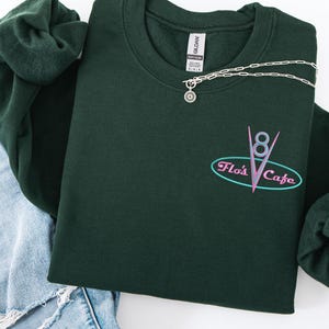 Puede incluir: Una sudadera verde oscuro con el texto "Flo's Cafe" en un dise&ntilde;o retro. Un collar plateado con un colgante de sol est&aacute; drapeado sobre la sudadera. La sudadera se combina con unos vaqueros azules y una goma de pelo verde.