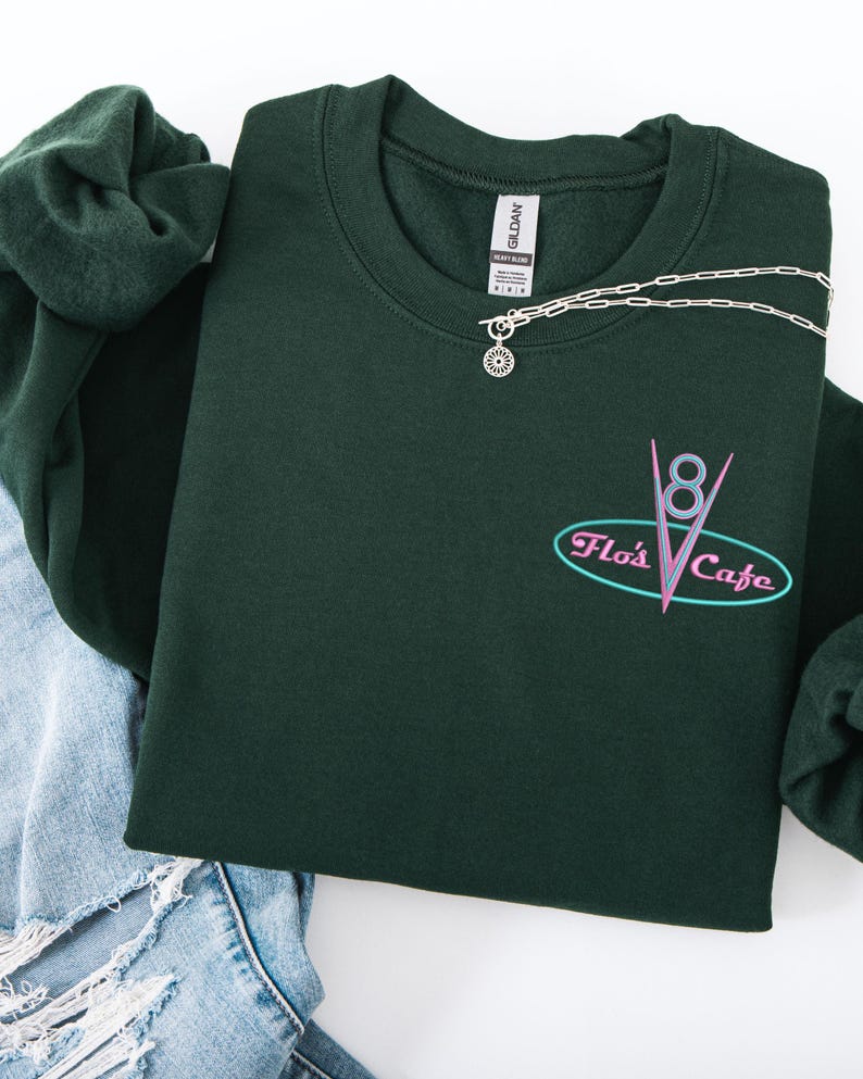 Puede incluir: Una sudadera verde oscuro con el texto "Flo's Cafe" en un dise&ntilde;o retro. Un collar plateado con un colgante de sol est&aacute; drapeado sobre la sudadera. La sudadera se combina con unos vaqueros azules y una goma de pelo verde.