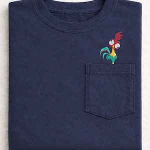 May include: Navy blue t-shirt with a pocket featuring an embroidered cartoon rooster. The rooster has red, yellow, and teal accents. The shirt is folded neatly, showcasing the design on the pocket.