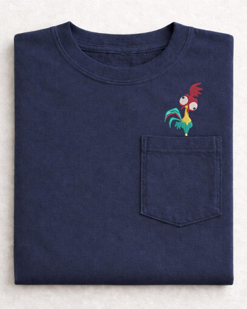 May include: Navy blue t-shirt with a pocket featuring an embroidered cartoon rooster. The rooster has red, yellow, and teal accents. The shirt is folded neatly, showcasing the design on the pocket.