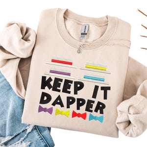 May include: A beige sweatshirt with the text "KEEP IT DAPPER" in black. The design includes colorful bow ties and stacks of colorful rectangles. A silver necklace with a pendant is visible. The sweatshirt is folded on top of a pair of blue jeans.