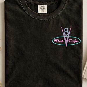Puede incluir: Camiseta negra con un logotipo de estilo ne&oacute;n que dice "Flo's Cafe". El logotipo presenta un "8" estilizado y una forma de "V", todo dentro de un marco ovalado. La camiseta tiene un cuello redondo cl&aacute;sico y una peque&ntilde;a etiqueta cerca del cuello.