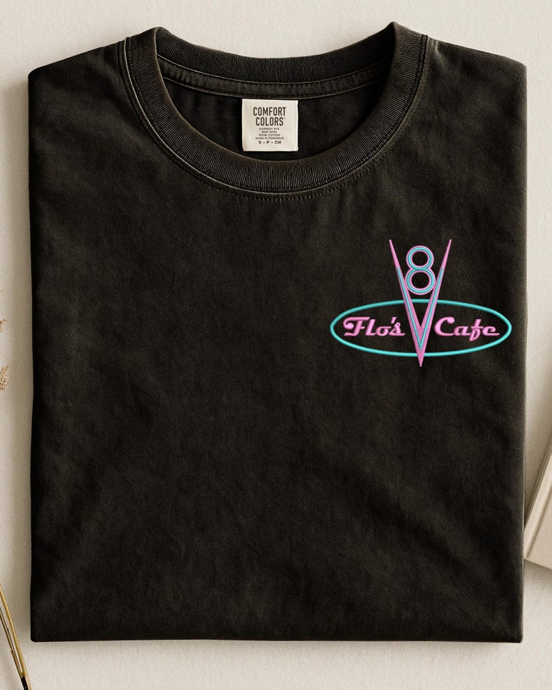 Puede incluir: Camiseta negra con un logotipo de estilo ne&oacute;n que dice "Flo's Cafe". El logotipo presenta un "8" estilizado y una forma de "V", todo dentro de un marco ovalado. La camiseta tiene un cuello redondo cl&aacute;sico y una peque&ntilde;a etiqueta cerca del cuello.