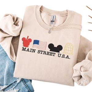 K&ouml;nnte beinhalten: Beiges Sweatshirt mit Rundhalsausschnitt und dem schwarzen Schriftzug "MAIN STREET U.S.A.", mit roten, blauen und schwarzen Grafiken. Eine silberne Halskette mit kleinem Anh&auml;nger ist sichtbar. Das Sweatshirt liegt auf einer blauen Jeans.