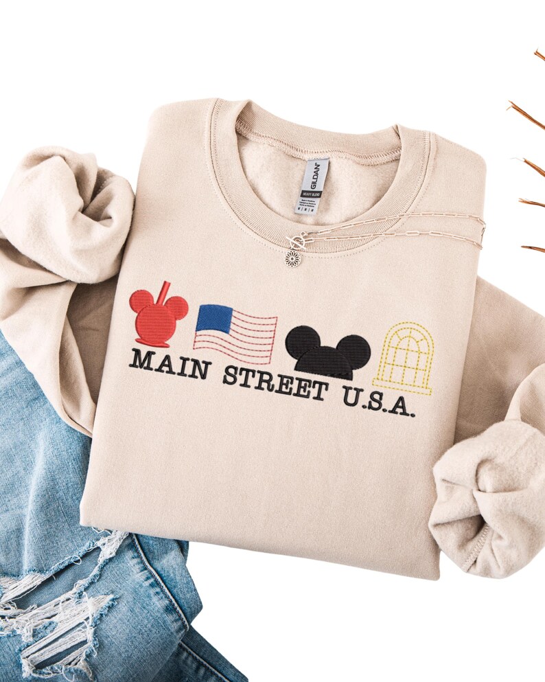 K&ouml;nnte beinhalten: Beiges Sweatshirt mit Rundhalsausschnitt und dem schwarzen Schriftzug "MAIN STREET U.S.A.", mit roten, blauen und schwarzen Grafiken. Eine silberne Halskette mit kleinem Anh&auml;nger ist sichtbar. Das Sweatshirt liegt auf einer blauen Jeans.