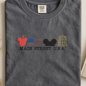 K&ouml;nnte beinhalten: Graues T-Shirt mit gestickten Designs: ein roter Mickey Mouse-Kopf, eine blaue Flagge, ein schwarzer Mickey Mouse-Kopf und ein goldener Bogenfenster. Der Text "MAIN STREET U.S.A." ist darunter gestickt. Das Shirt hat einen Rundhalsausschnitt und ein "COMFORT COLORS"-Etikett.