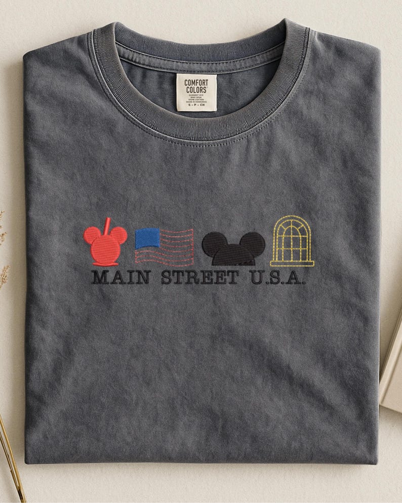 K&ouml;nnte beinhalten: Graues T-Shirt mit gestickten Designs: ein roter Mickey Mouse-Kopf, eine blaue Flagge, ein schwarzer Mickey Mouse-Kopf und ein goldener Bogenfenster. Der Text "MAIN STREET U.S.A." ist darunter gestickt. Das Shirt hat einen Rundhalsausschnitt und ein "COMFORT COLORS"-Etikett.