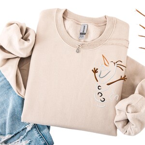May include: A beige crewneck sweatshirt with an embroidered Olaf design. The design includes a carrot nose, twig arms, and three black buttons. A silver necklace with a pendant is on the sweatshirt. The sweatshirt is folded on top of blue jeans.