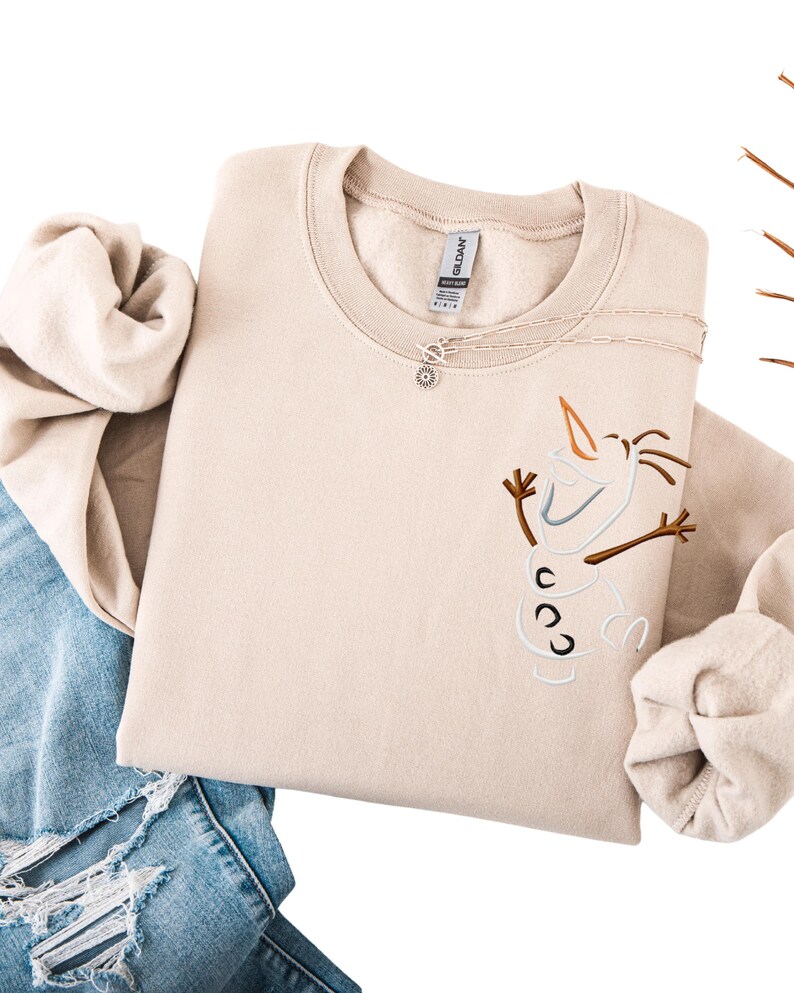 May include: A beige crewneck sweatshirt with an embroidered Olaf design. The design includes a carrot nose, twig arms, and three black buttons. A silver necklace with a pendant is on the sweatshirt. The sweatshirt is folded on top of blue jeans.