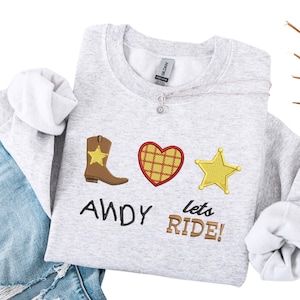 Puede incluir: Una sudadera gris claro con gráficos bordados con temática de vaquero. El diseño incluye una bota marrón con una estrella amarilla, un corazón rojo y una estrella de sheriff amarilla. El texto dice "ANDY lets RIDE!"