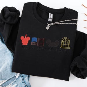 K&ouml;nnte beinhalten: Schwarzes Sweatshirt mit Rundhalsausschnitt und gesticktem Main Street U.S.A.-Design. Mit rotem Mickey Mouse-Kopf, amerikanischer Flagge, schwarzer Mickey Mouse-Silhouette und gelbem Fenster. Eine silberne Halskette mit rundem Anh&auml;nger liegt auf dem Sweatshirt.