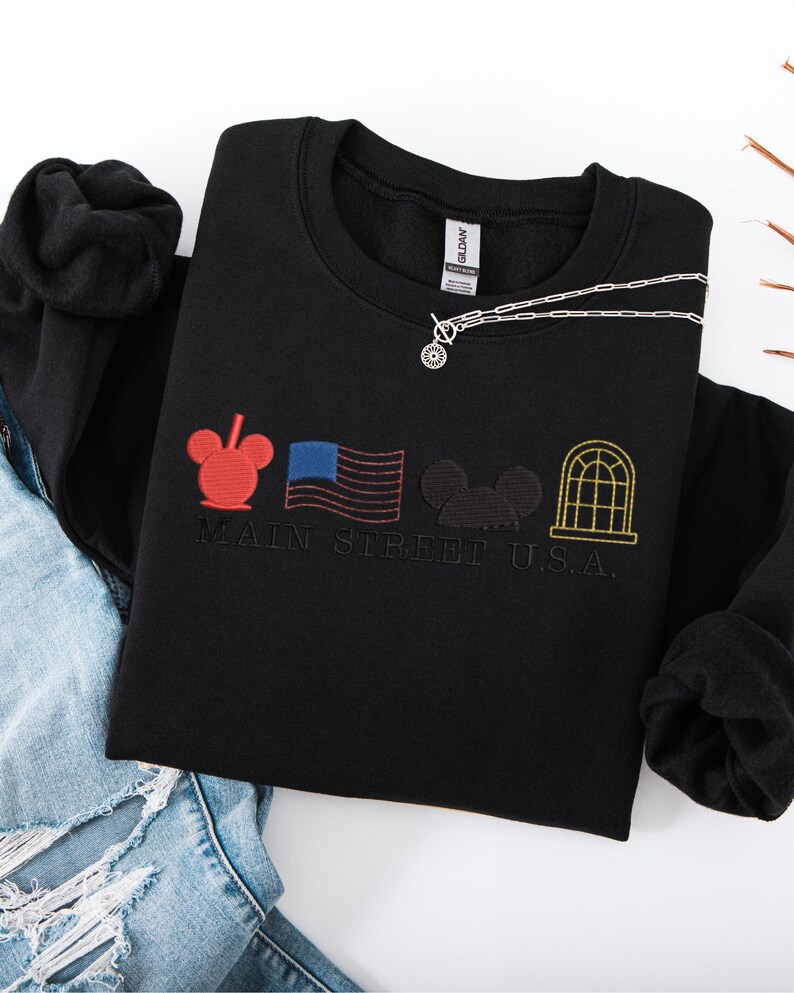 K&ouml;nnte beinhalten: Schwarzes Sweatshirt mit Rundhalsausschnitt und gesticktem Main Street U.S.A.-Design. Mit rotem Mickey Mouse-Kopf, amerikanischer Flagge, schwarzer Mickey Mouse-Silhouette und gelbem Fenster. Eine silberne Halskette mit rundem Anh&auml;nger liegt auf dem Sweatshirt.