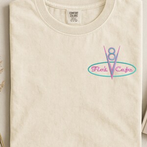 Puede incluir: Camiseta color crema con cuello redondo. Presenta un gr&aacute;fico de estilo ne&oacute;n de un emblema "V8" con el texto "Flo's Cafe" en un &oacute;valo turquesa. La camiseta tiene una etiqueta que dice "Comfort Colors".