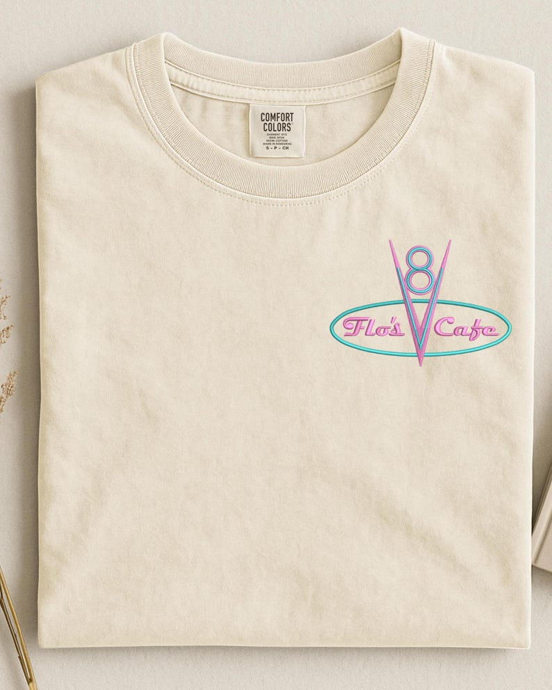 Puede incluir: Camiseta color crema con cuello redondo. Presenta un gr&aacute;fico de estilo ne&oacute;n de un emblema "V8" con el texto "Flo's Cafe" en un &oacute;valo turquesa. La camiseta tiene una etiqueta que dice "Comfort Colors".