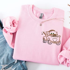 Puede incluir: Una sudadera rosa claro con un dise&ntilde;o bordado de un cami&oacute;n de remolque de dibujos animados. La sudadera de cuello redondo tiene mangas largas y se combina con vaqueros azules. Un delicado collar con un peque&ntilde;o colgante est&aacute; drapeado sobre la sudadera.