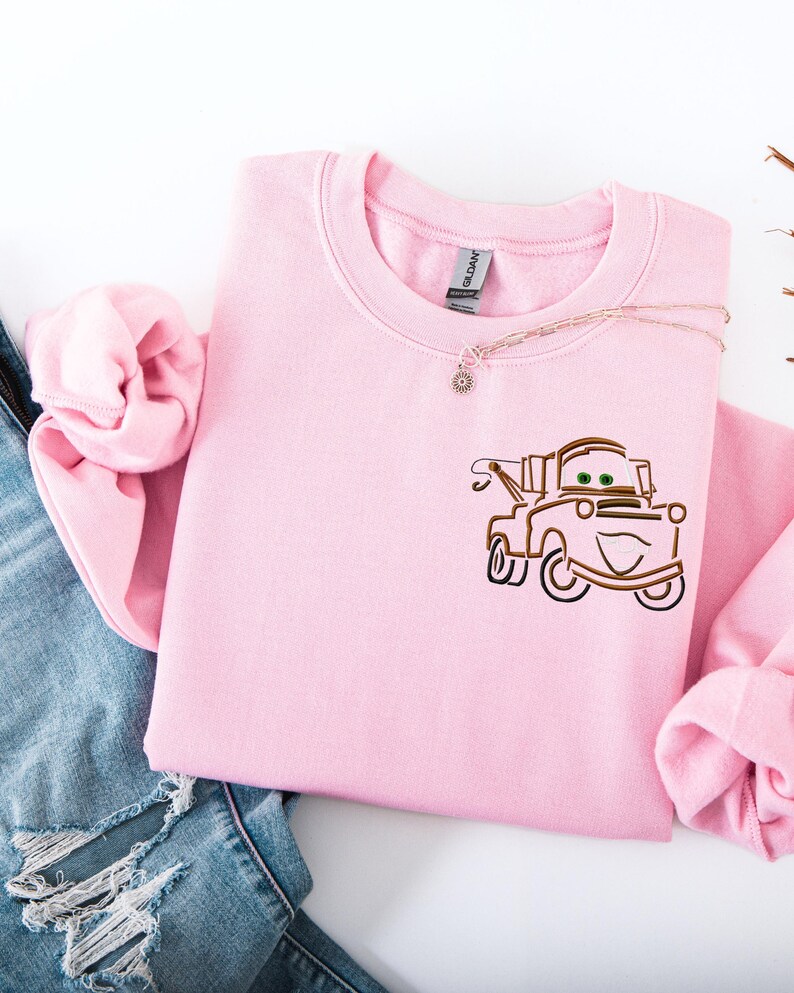 Puede incluir: Una sudadera rosa claro con un dise&ntilde;o bordado de un cami&oacute;n de remolque de dibujos animados. La sudadera de cuello redondo tiene mangas largas y se combina con vaqueros azules. Un delicado collar con un peque&ntilde;o colgante est&aacute; drapeado sobre la sudadera.