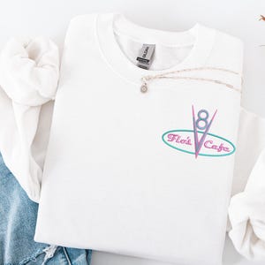 Puede incluir: Sudadera blanca de cuello redondo con el texto bordado "Flo's Cafe" en un dise&ntilde;o ovalado turquesa y rosa. Un collar plateado con un peque&ntilde;o colgante descansa sobre la sudadera. La sudadera est&aacute; doblada sobre una superficie blanca.