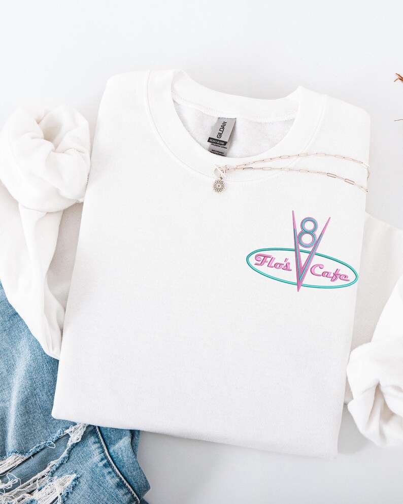 Puede incluir: Sudadera blanca de cuello redondo con el texto bordado "Flo's Cafe" en un dise&ntilde;o ovalado turquesa y rosa. Un collar plateado con un peque&ntilde;o colgante descansa sobre la sudadera. La sudadera est&aacute; doblada sobre una superficie blanca.