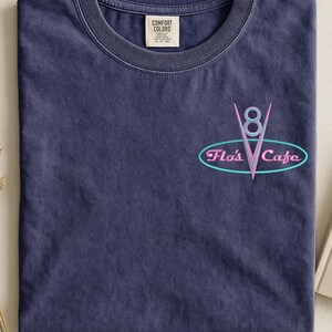 Puede incluir: Una camiseta azul marino con cuello redondo y un peque&ntilde;o gr&aacute;fico en la parte delantera. El gr&aacute;fico presenta un dise&ntilde;o de estilo ne&oacute;n del texto "Flo's Cafe" en un &oacute;valo turquesa, con un "8" rosa y una forma de "V" rosa.