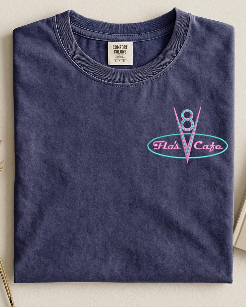 Puede incluir: Una camiseta azul marino con cuello redondo y un peque&ntilde;o gr&aacute;fico en la parte delantera. El gr&aacute;fico presenta un dise&ntilde;o de estilo ne&oacute;n del texto "Flo's Cafe" en un &oacute;valo turquesa, con un "8" rosa y una forma de "V" rosa.