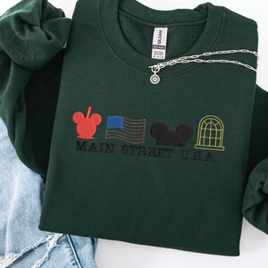 K&ouml;nnte beinhalten: Dunkelgr&uuml;nes Sweatshirt mit gesticktem Main Street U.S.A. Design. Es zeigt einen roten Mickey Mouse-Kopf, eine blaue amerikanische Flagge, einen schwarzen Mickey Mouse-Kopf und ein gelbes Fenster. Eine silberne Halskette mit Sonnenanh&auml;nger liegt darauf.